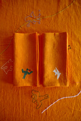 Two folded orange napkins with embroidered designs on an orange background