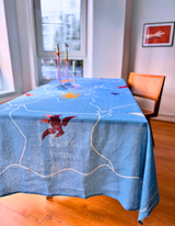Blue tablecloth with various coloured birds on a wooden table in a room with large windows.
