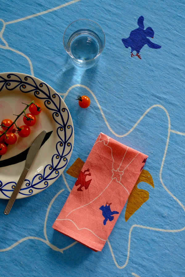 Table setting with a pink napkin featuring blue bear designs, a glass of blue liquid, and a plate with tomatoes and a knife on a blue tablecloth.