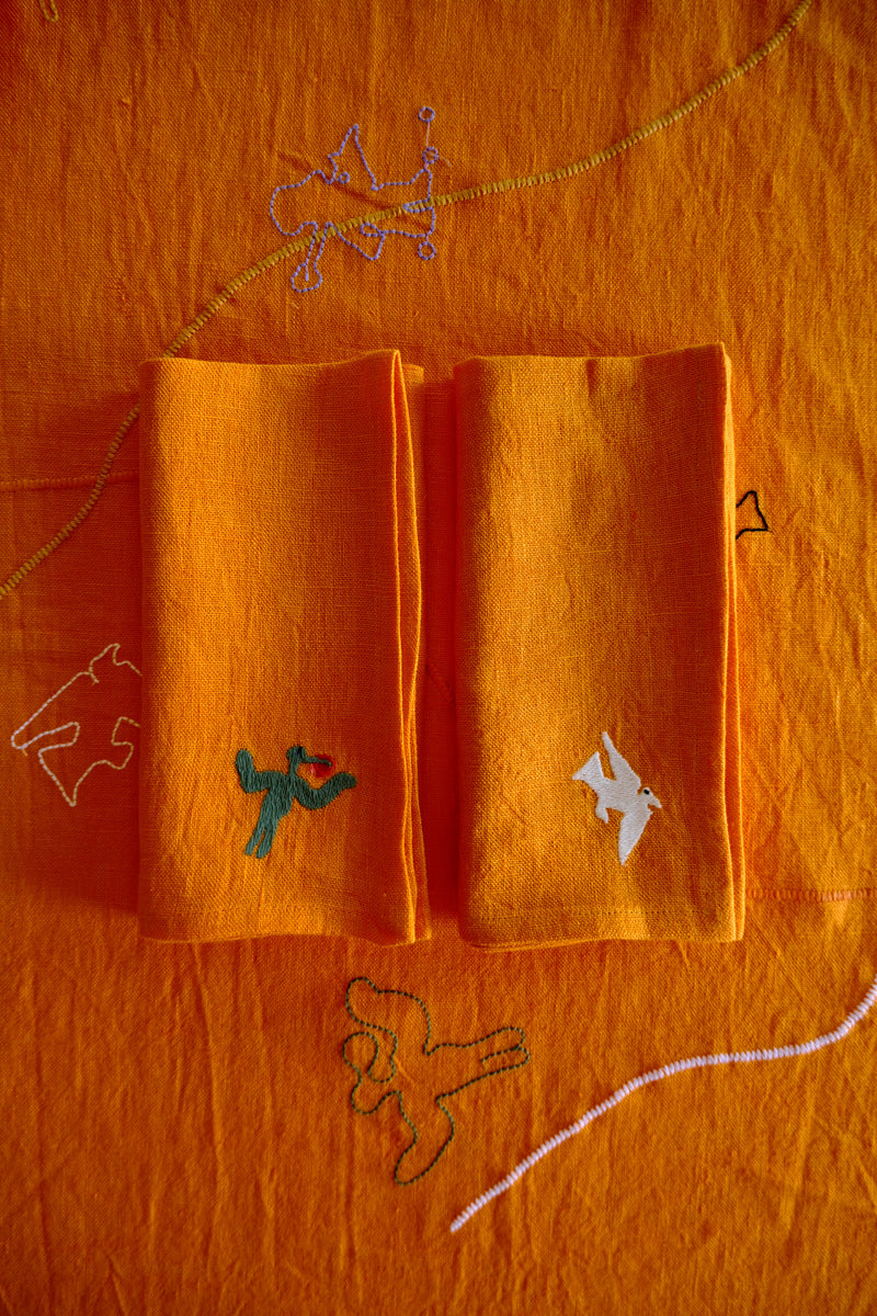 Two folded orange napkins with embroidered designs on an orange background