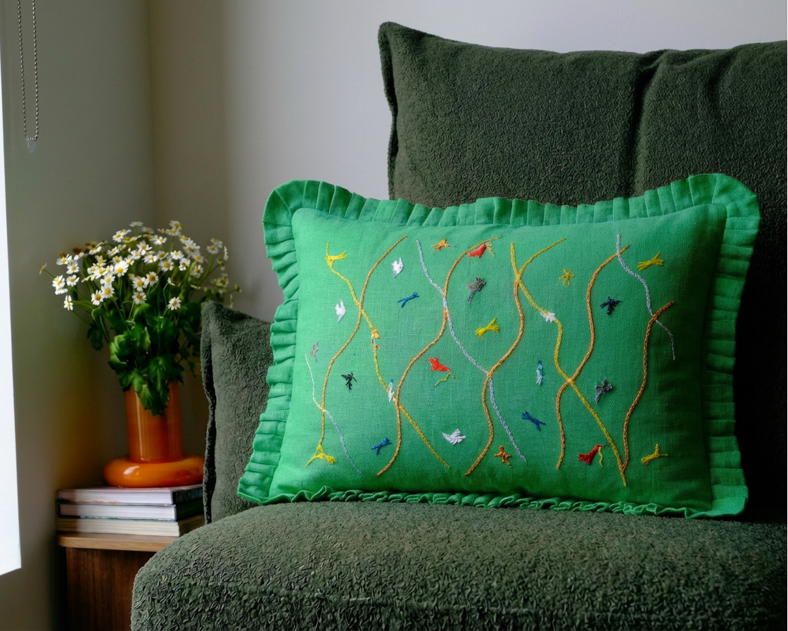 Green embroidered cushion on a green couch with a plant and books in the background.
